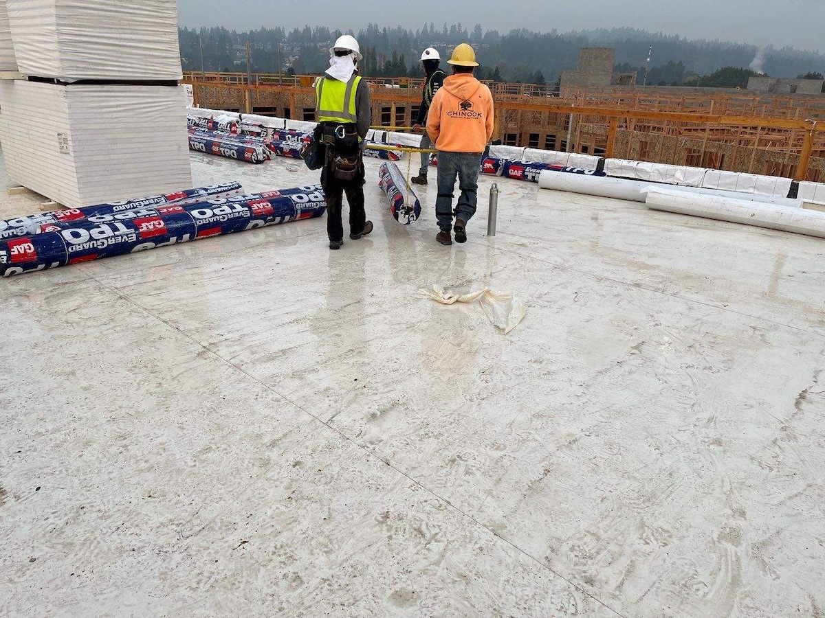 Three GAF contractors on a roof with rolls of GAF EverGuard TPO laying on the roof, while two GAF contractors carry a roll of GAF EverGuard TPO